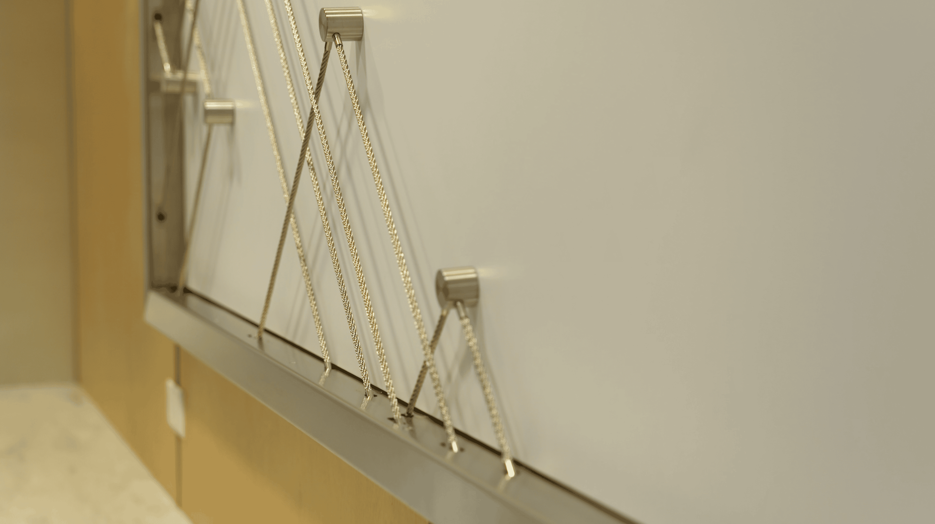 Close-up view of a modern wall installation featuring diagonal metal cables mounted on a white background. The cables are secured by brushed metal fixtures, forming an intricate geometric pattern. The installation is part of a facility branding design in an office setting, framed by wood and metal elements.